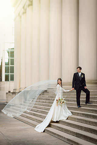Bride with veil flowing in the breeze and Groom on staircase with building pillars 